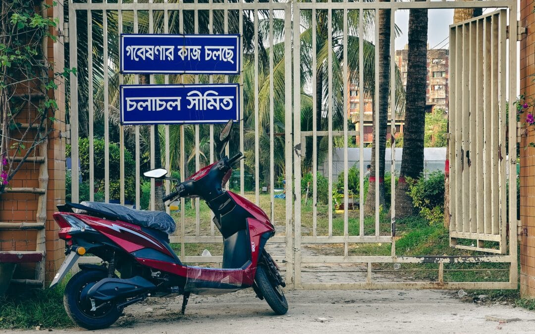 Electric Two-Wheelers in India illustration showing a maroon electric scooter parked outside a white gate with Bengali signs