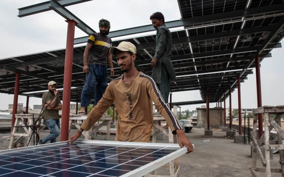 Local workers on a concrete rooftop in Pakistan installing solar panels on a large steel frame structure.