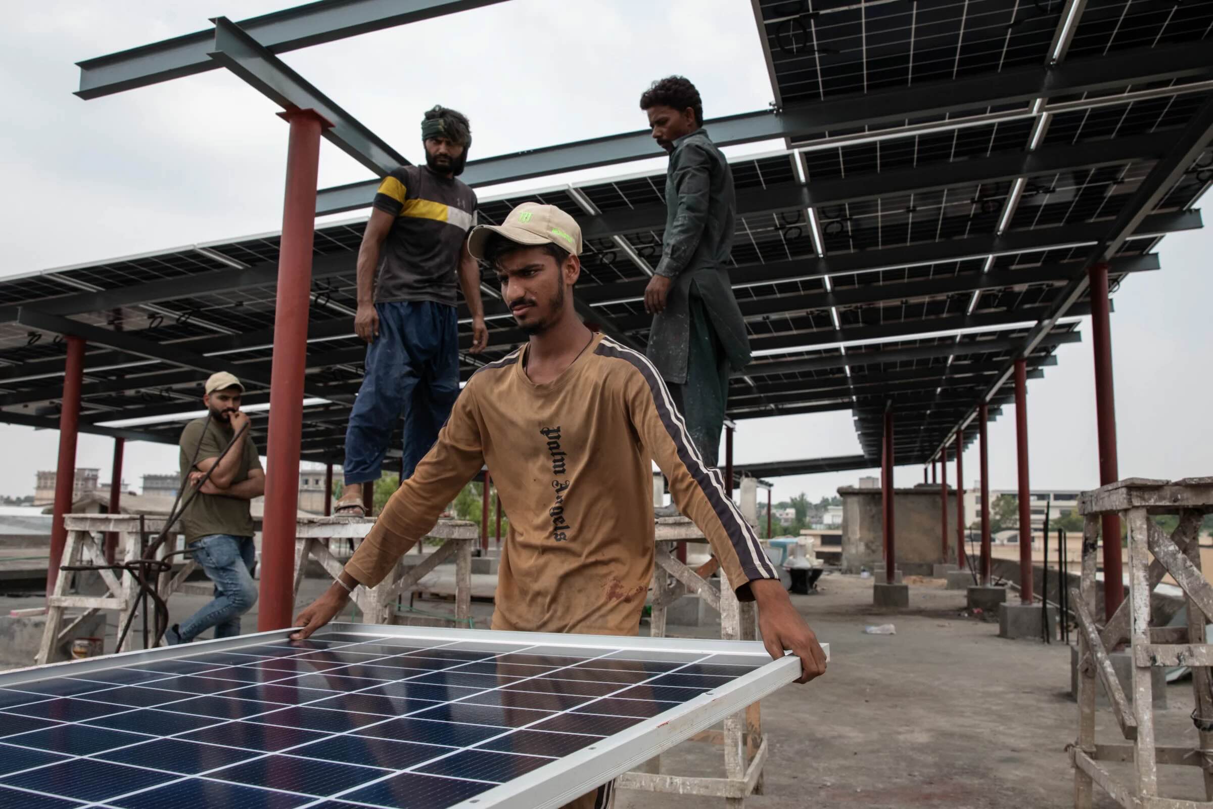 Local workers on a concrete rooftop in Pakistan installing solar panels on a large steel frame structure.